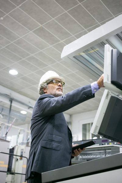 Serious mature male plant engineer operating industrial machine, pushing buttons on control panel, holding tablet. Low angle, vertical shot. Production process or machinery concept